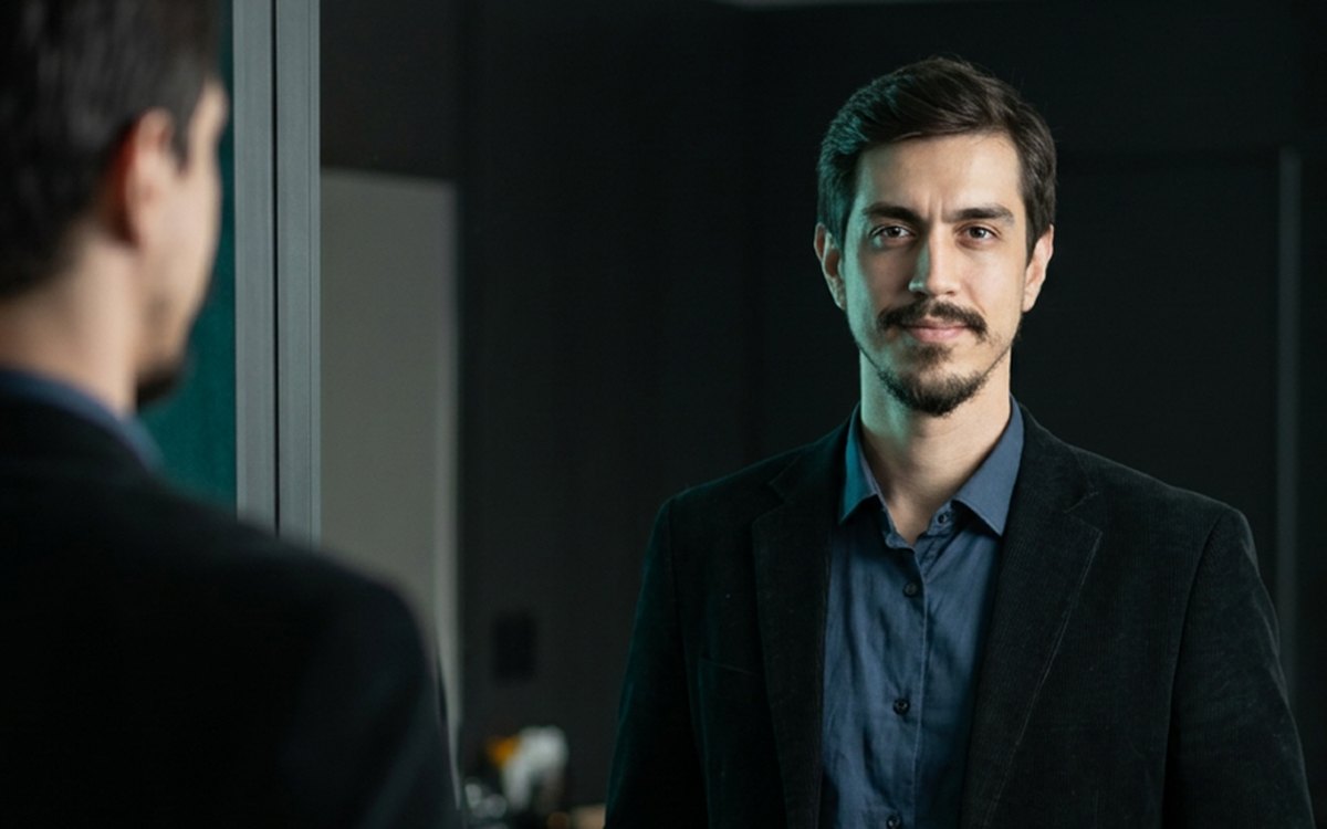 Homem de terno diante do proprio reflexo, ajustando a postura.