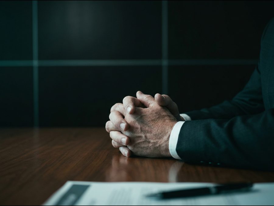 Maos entrelaçadas e tensas sobre a mesa, diante de um contrato e uma caneta.